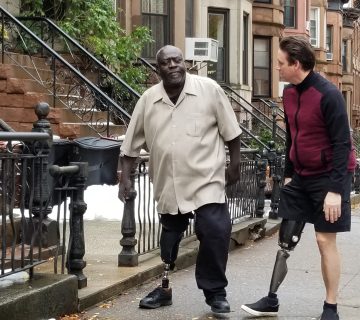 Two men, both with prosthetic legs, walk and converse on a sidewalk in a residential area with brownstone buildings