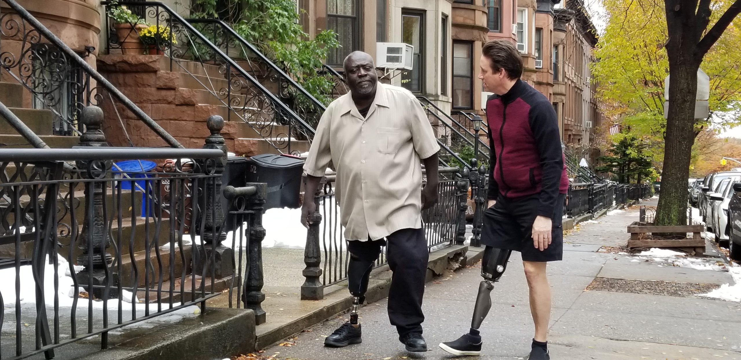 Two men, both with prosthetic legs, walk and converse on a sidewalk in a residential area with brownstone buildings