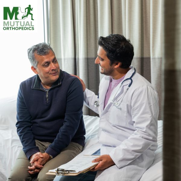 A doctor in a white coat discusses amputation rehabilitation with a seated older man, holding a medical chart. They are in a room with curtains, focused on recovery strategies. The logo in the corner reads "Mutual Orthopedics," symbolizing care and expertise in Islandia. A doctor in a white coat discusses amputation rehabilitation with a seated older man, holding a medical chart They are in a room with curtains, focused on recovery strategies The logo in the corner reads "Mutual Orthopedics," symbolizing care and expertise in Islandia
