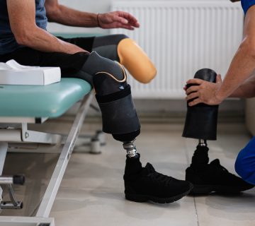 A healthcare worker helps a person with two prosthetic legs adjust their right prosthesis while they sit on a medical examination table