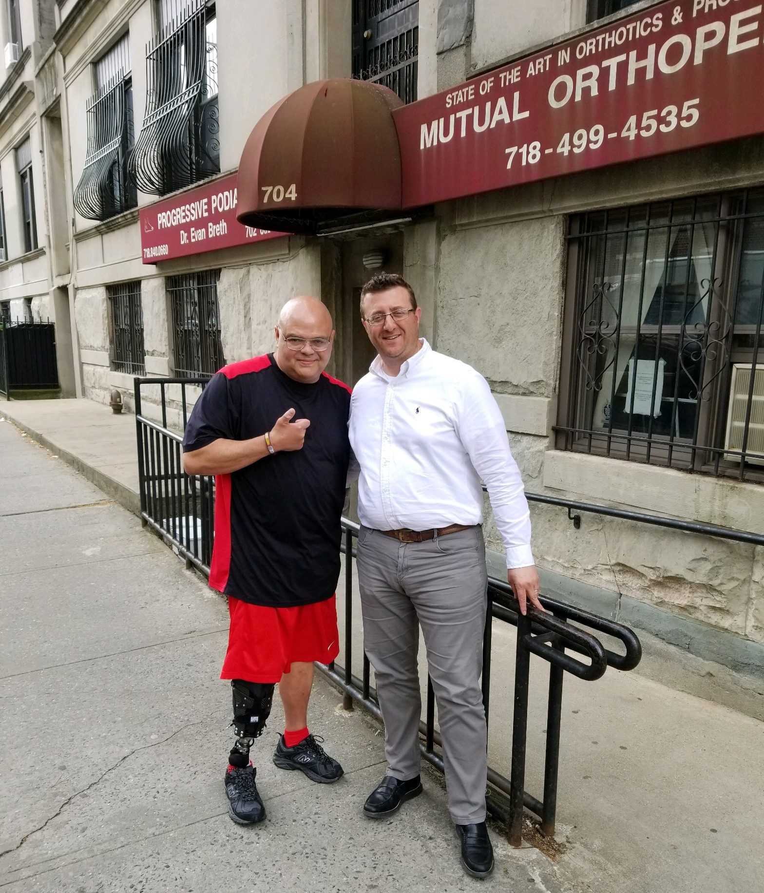 Two men stand and pose for a photo on a sidewalk in front of a building with a "Mutual Orthopedic" sign; one man has a prosthetic leg 