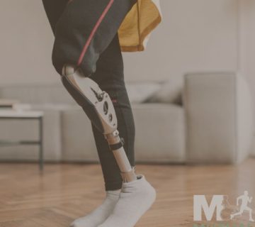 A person with a prosthetic leg stands on a wooden floor in a living room, wearing black pants and white socks The Mutual Orthopedics logo is visible in the corner